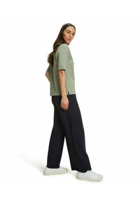 Olijfgroene top met korte mouwen gecombineerd met marineblauwe wijde broek en witte sneakers, die een ontspannen pasvorm en zachte stoftextuur tonen.