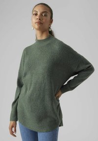 Green knitted sweater with a high neck, loose fit, ribbed cuffs, and hem, paired with blue denim jeans. Soft texture, minimal design.