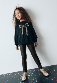 Sudadera negra con detalle de lazo de lentejuelas, leggings negros y zapatos Mary Jane dorados, sobre un suelo cubierto de confeti.