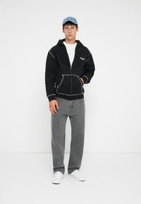 Svart hettejakke med hvite sømmer, frontlommer og logo på brystet, paret med grå jeans og hvite sneakers. Denim caps på hodet.