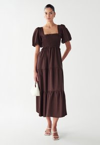 Robe maxi brune à manches bouffantes avec un corsage smocké et une jupe à volants. Associée à des sandales blanches à lanières et à un petit sac blanc.