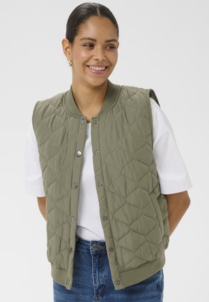 Femme souriante portant un gilet matelassé vert olive sur un t-shirt blanc et un jean bleu, debout les mains derrière le dos.