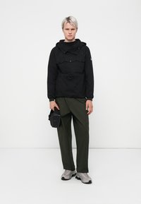 Black pullover jacket with a drawstring waist and two chest pockets, paired with olive green trousers and gray sneakers. Small black bag attached.