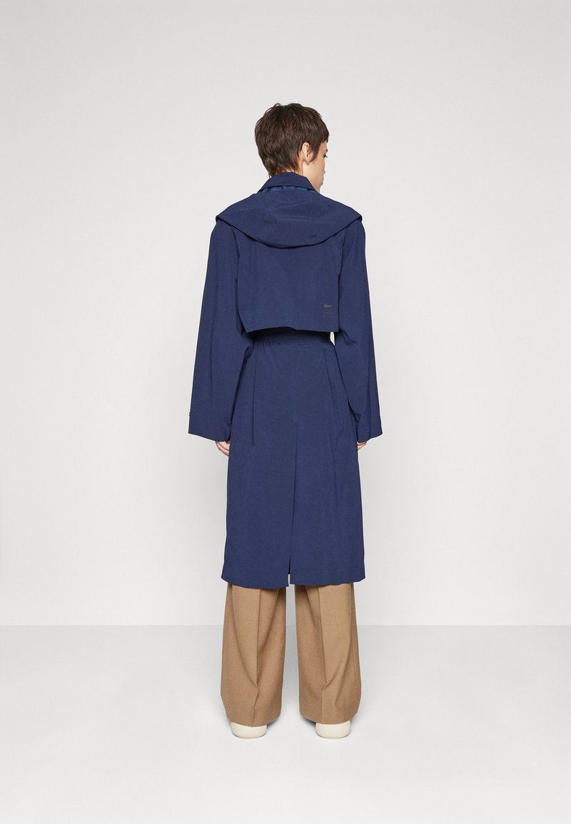 Manteau trench bleu marine avec capuche, manches longues, taille cintrée et fente au dos, fabriqué en tissu lisse, avec un logo minimal.