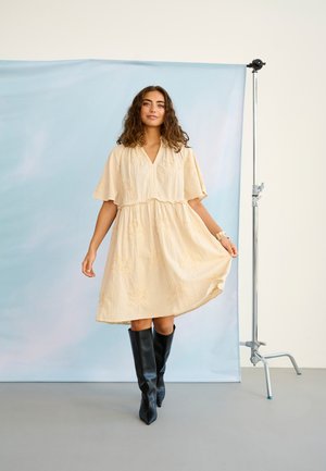 Woman in a cream knee-length dress with embroidery and black knee-high boots stands on gray floor against blue backdrop.