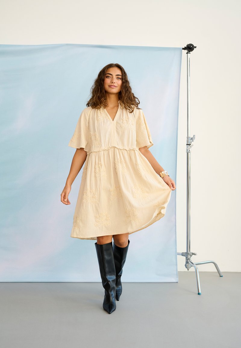 Femme en robe crème longueur genou avec broderies et bottes noires montant jusqu'aux genoux, debout sur un sol gris devant un fond bleu.