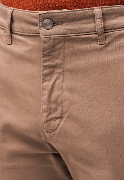 Gros plan d'une personne portant un pantalon marron clair avec un bouton et une fermeture éclair, associé à un haut tricoté orange.