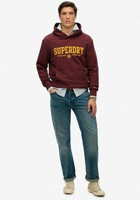 Felpa maroon con logo "SUPERDRY" giallo, camicia a quadri azzurri sotto, jeans blu e sneakers beige. Look casual.
