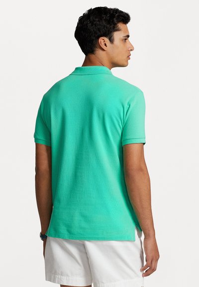 Polo en turquoise en tissu respirant, à manches courtes, col côtelé, fentes latérales au bas. Porté avec un short blanc, présenté de dos.