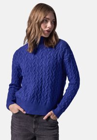 Pull en tricot bleu avec un col montant et des poignets côtelés, présentant un motif texturé. Associé à un pantalon gris foncé.