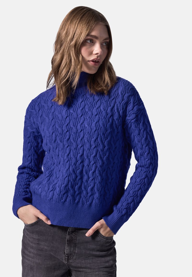 Pull en tricot bleu avec un col montant et des poignets côtelés, présentant un motif texturé. Associé à un pantalon gris foncé.