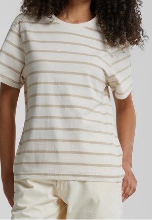 Osoba ubrana w beżowo-biały t-shirt w poziome paski na krótki rękaw oraz jasne spodnie, na jednolitym tle.