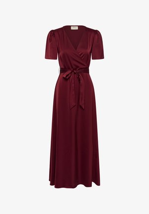 Femme aux cheveux longs portant une robe portefeuille bordeaux avec manches bouffantes, des sandales à talons noires, des boucles d'oreilles créoles dorées et un collier sur un fond uni.