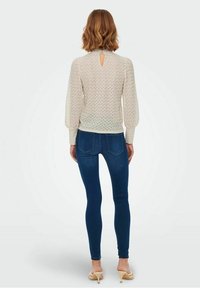 Blusa di tessuto crema con maniche a sbuffo e dettaglio a forma di chiave sul retro, abbinata a jeans skinny blu scuro e tacchi di colore chiaro.