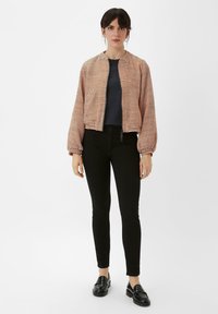 Rote und cremefarbene Bomberjacke mit Muster, mit einem Reißverschluss vorne, weit geschnittenen Ärmeln, zwei Seitentaschen, kombiniert mit schwarzen Skinny-Jeans und Slippern.