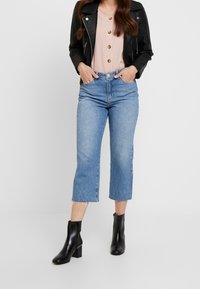 Jeans cropped in denim chiaro con orli sfrangiati, abbinati a una camicia rosa con bottoni e a una giacca di pelle nera. Stivaletti neri con tacco.