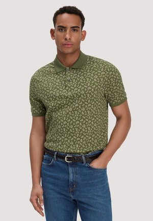 Mann trägt grünes kurzärmeliges Poloshirt mit Blumenmuster, in blaue Jeans gesteckt, mit schwarzem geflochtenem Gürtel, steht mit einer Hand in der Tasche.