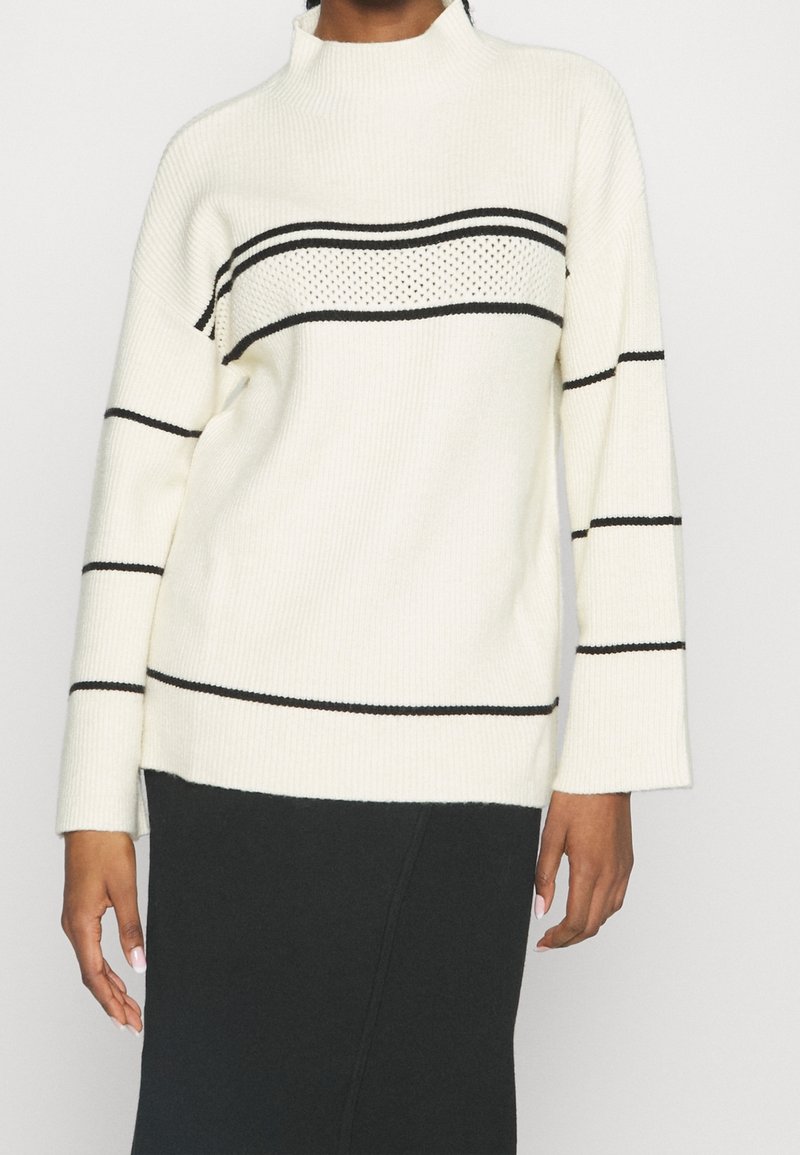 Vit stickad tröja med svarta horisontella ränder. Har hög hals och ribbad textur. Långa ärmar och avslappnad passform.