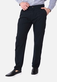 Pantaloni in cotone di colore blu navy con una vestibilità sartoriale, dotati di chiusura a bottone e tasche laterali. Abbinati a scarpe slip-on nere su uno sfondo bianco.