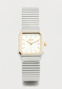 Ασημένιο και χρυσό ρολόι χειρός Lacoste με ορθογώνιο καντράν, χρυσούς δείκτες ώρας και ευέλικτο μεταλλικό λουράκι σε λευκό φόντο.