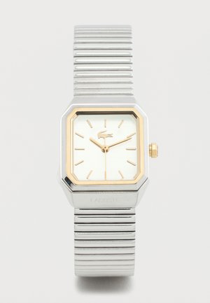 Ezüst és arany tónusú Lacoste karóra téglalap alakú számlappal, arany óraszámozással és rugalmas fémszíjjal, fehér háttéren.