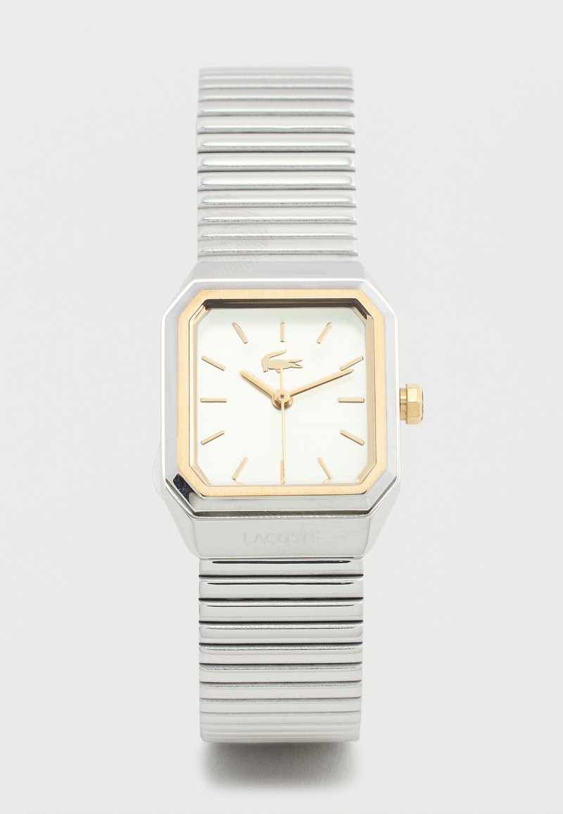Ασημένιο και χρυσό ρολόι χειρός Lacoste με ορθογώνιο καντράν, χρυσούς δείκτες ώρας και ευέλικτο μεταλλικό λουράκι σε λευκό φόντο.