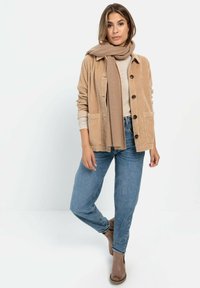 Beige Cord-Jacket mit Knöpfen, braunem Schal, beigem Oberteil, blauen Jeans und braunen Stiefeletten. Hat zwei Fronttaschen.