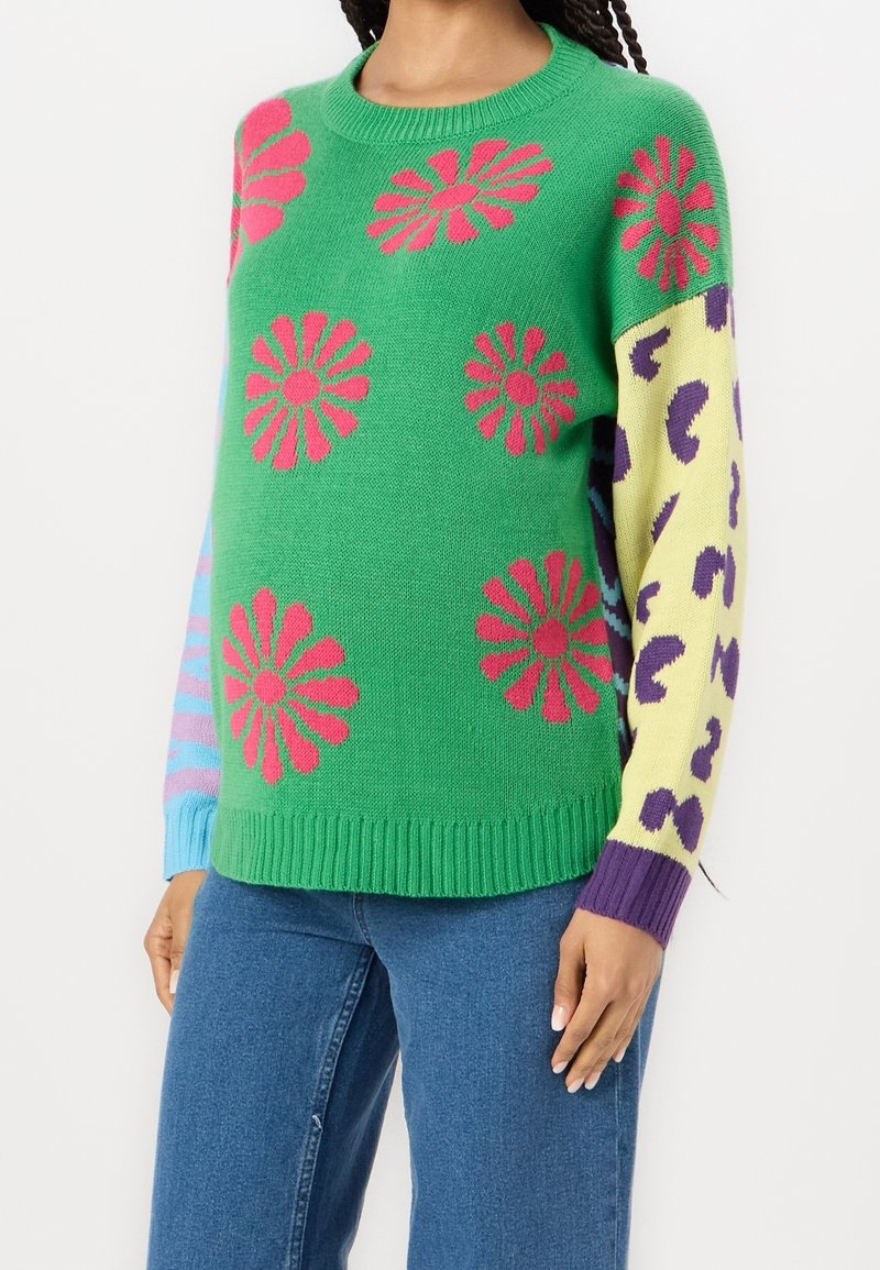 Femme portant un pull vert avec des fleurs roses, une manche jaune avec des taches léopard violettes, et une manche bleue avec des rayures roses, associée à un jean bleu.