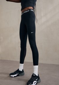 Sorte Nike Pro leggings med tætsiddende pasform, der har et hvidt Nike-logo på låret. Båret med sorte sneakers og hvide sokker.