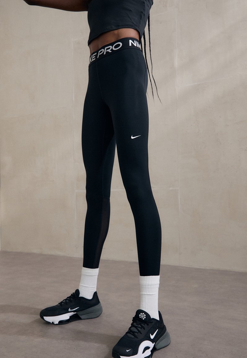 Sorte Nike Pro leggings med tætsiddende pasform, der har et hvidt Nike-logo på låret. Båret med sorte sneakers og hvide sokker.