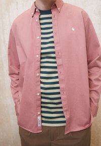 Helles rosa Hemd mit Knopfleiste über einem grün- und navygestreiften T-Shirt. Es hat einen klassischen Kragen und weiße Knöpfe, mit einem Etikett am Saum.