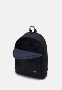 Mochila negra con un exterior suave, cierre con cremallera y un bolsillo interior en color azul marino. Cuenta con una parte superior redondeada y un pequeño logo en el frente.