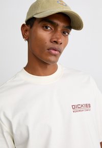 T-shirt color crema con colletto rotondo, caratterizzata da un logo rosso scuro. Il modello indossa un cappellino verde chiaro con una toppa ricamata.
