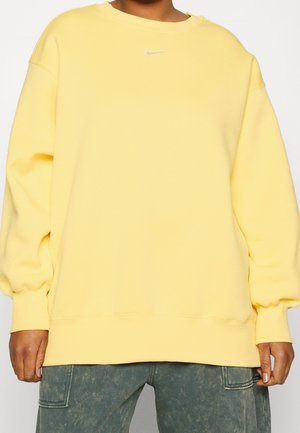 Person trägt ein lockeres, pastellgelbes Nike-Sweatshirt kombiniert mit ausgewaschenen grünen Cargohosen und steht vor einem schlichten Hintergrund.