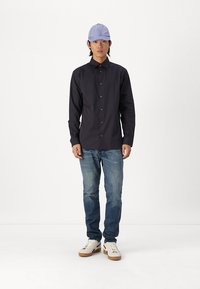 Black striped button-up shirt, light blue cap, denim jeans, and white shoes with brown soles. Model stands against a plain background.