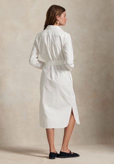 Nő hosszú ujjú, öves, fehér ingruha, oldalsó hasítékkal, oldalra állva, fekete lapos cipőben és lógó fülbevalóval.