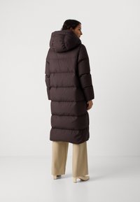 Manteau matelassé long de couleur marron foncé, présentant une capuche, une coupe oversize, des sections matelassées horizontales et des zips latéraux, associé à un pantalon beige.
