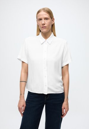 Camisa branca de manga curta com botões, gola simples e tecido liso, combinada com jeans azul escuro. A camisa tem um corte descontraído.