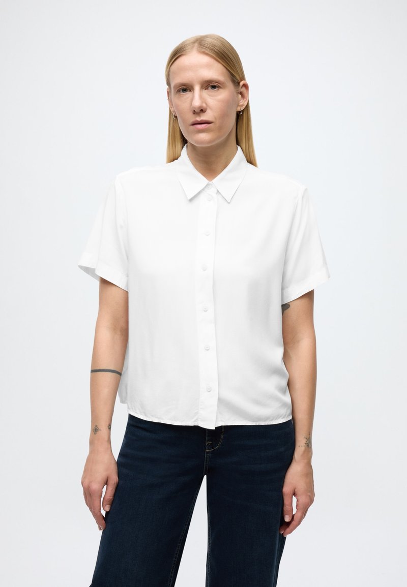 Fehér rövid ujjú, gombos ing egyszerű gallérral és sima anyagból, sötétkék farmerral párosítva. Az ing laza fazonú.