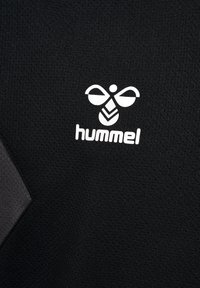Czarna tkana tkanina z teksturowanym splotem, z białym logo 'hummel' i wzorem pszczoły nadrukowanym na powierzchni. Podkreślona ciemnoszarym kształtem geometrycznym.