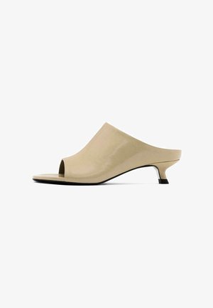 Beige open-toe mules with a sleek, glossy finish and a low, angular heel. The design features smooth curves and a minimalist silhouette.