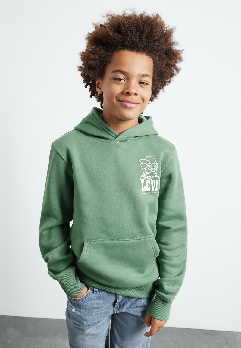 Felpa verde con tasca frontale e polsini a coste, caratterizzata da un logo grafico bianco sul petto. Indossata con jeans chiari strappati.