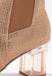 Zapato de tacón alto beige con superficie texturizada y tachuelas doradas, que cuenta con un tacón de bloque transparente y un panel de tela en contraste.