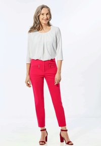 Haut blanc à manches trois-quarts associé à un pantalon slim fit rose vif avec des zips sur les côtés. Le modèle porte des sandales à talons carrés rouges.
