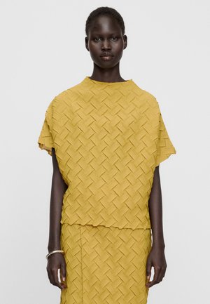Vrouw staat frontaal, draagt een gele getextureerde top met korte mouwen en een rok met geometrisch patroon tegen een effen lichte achtergrond.