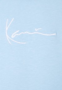 Białe, kursywą wyszyte logo "Kamik" na jasnoniebieskim dzianinowym materiale, pokazujące szczegóły marki na tkaninie odzieżowej.