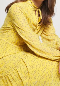 Vestido de estampado floral amarillo con escote volante y mangas largas. Presenta un detalle de lazo en el cuello y volantes en capas en el dobladillo.