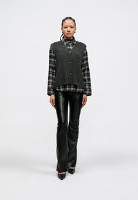 Γκρι πλεκτό γιλέκο με V-λαιμόκοψη και κουμπιά πάνω από μια ασπρόμαυρη καρό πουκάμισο, σε συνδυασμό με γυαλιστερά μαύρα δερμάτινα παντελόνια με καμπάνα.