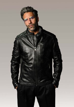 Homme aux cheveux et à la barbe grisonnants, portant des lunettes et une veste en cuir noire zippée, debout les mains dans les poches devant un fond uni.