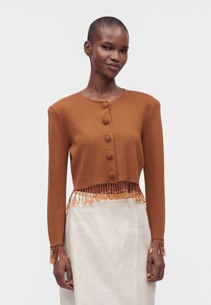 Kvinde iført en rustfarvet, knappet cropped cardigan med frynser og perledetaljer på manchetter og kant, stylet med et lyst beige skørt.